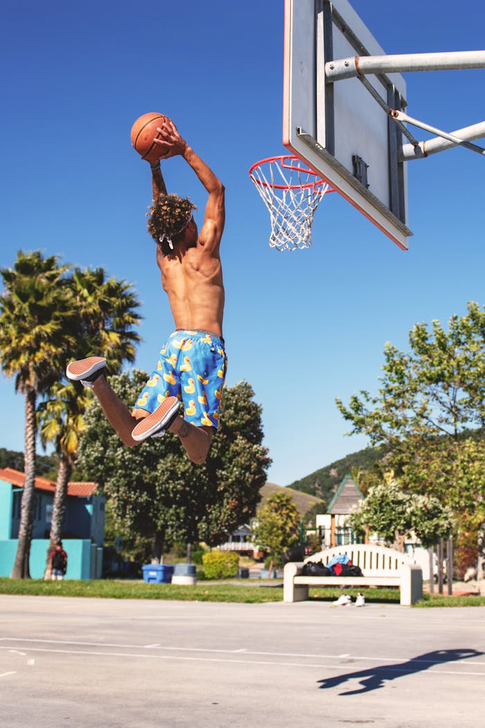 Shirtless man performing a basketball dunk at outdoor court in summer.